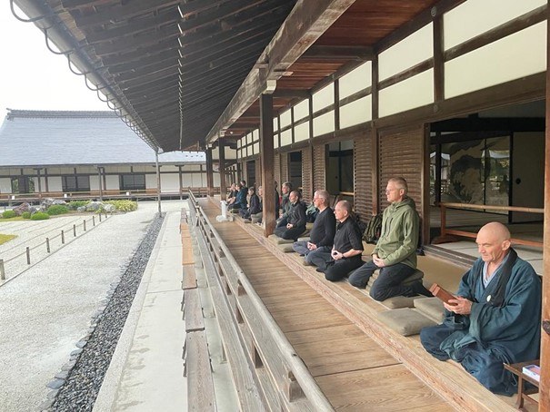 Op de veranda van een klooster in Kyoto zitten mensen te mediteren. Op de voorgrond zit een monnik met houten kleppers.